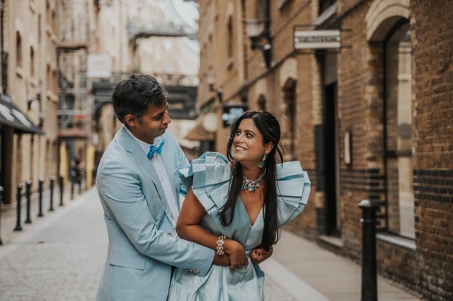 A smiling couple in pale blue clothes standing close with brick buildings softly behind them during their London family photoshoot.
