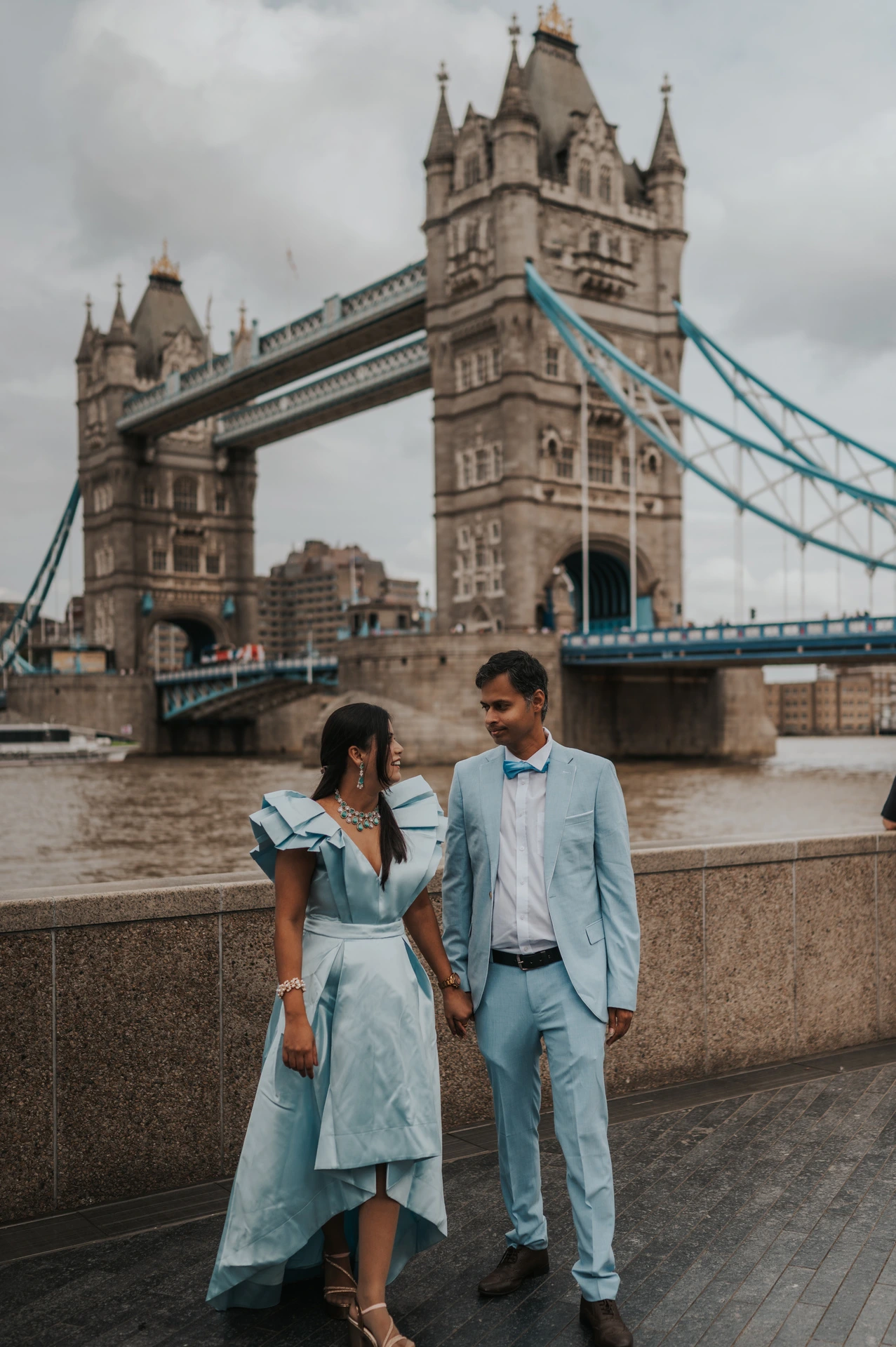 A couple holding hands while standing apart beside the riverside wall below Tower Bridge during their London family photoshoot.