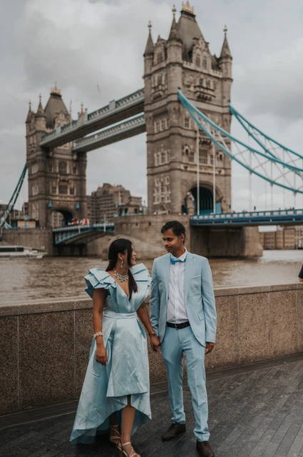 London family photoshoot: holding hands with the bridge behind them A couple holding hands while standing apart beside the riverside wall below Tower Bridge during their London family photoshoot.