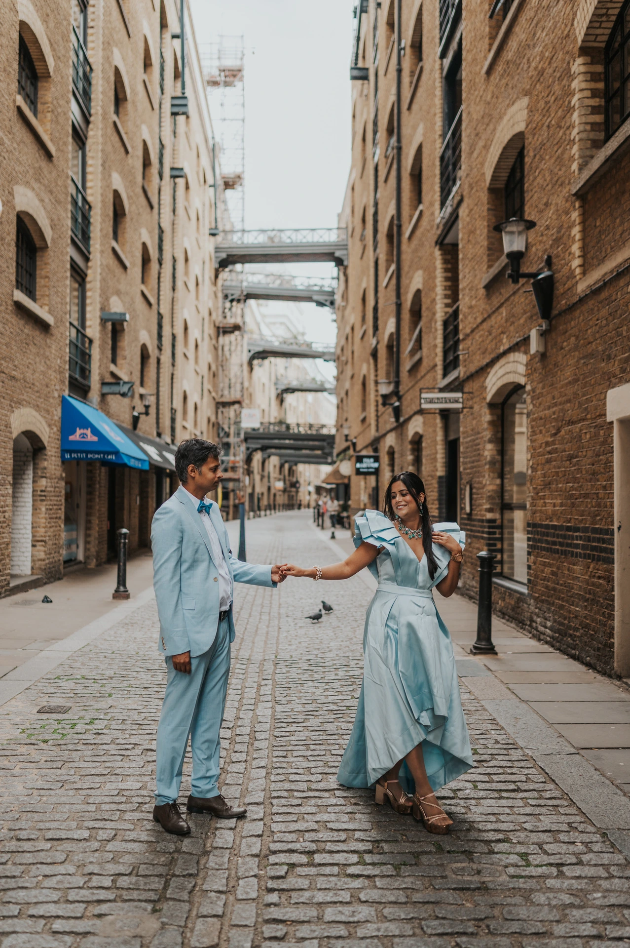 A couple holding hands as the woman turns in a pale blue dress in a cobbled lane during their London family photoshoot.