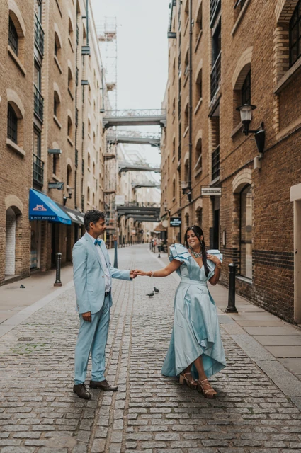 A couple holding hands as the woman turns in a pale blue dress in a cobbled lane during their London family photoshoot.