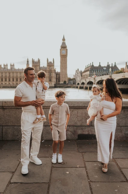 London family photoshoot: parents and children beside the river Parents holding their youngest children while an older child stands between them beside the Thames during their London family photoshoot.
