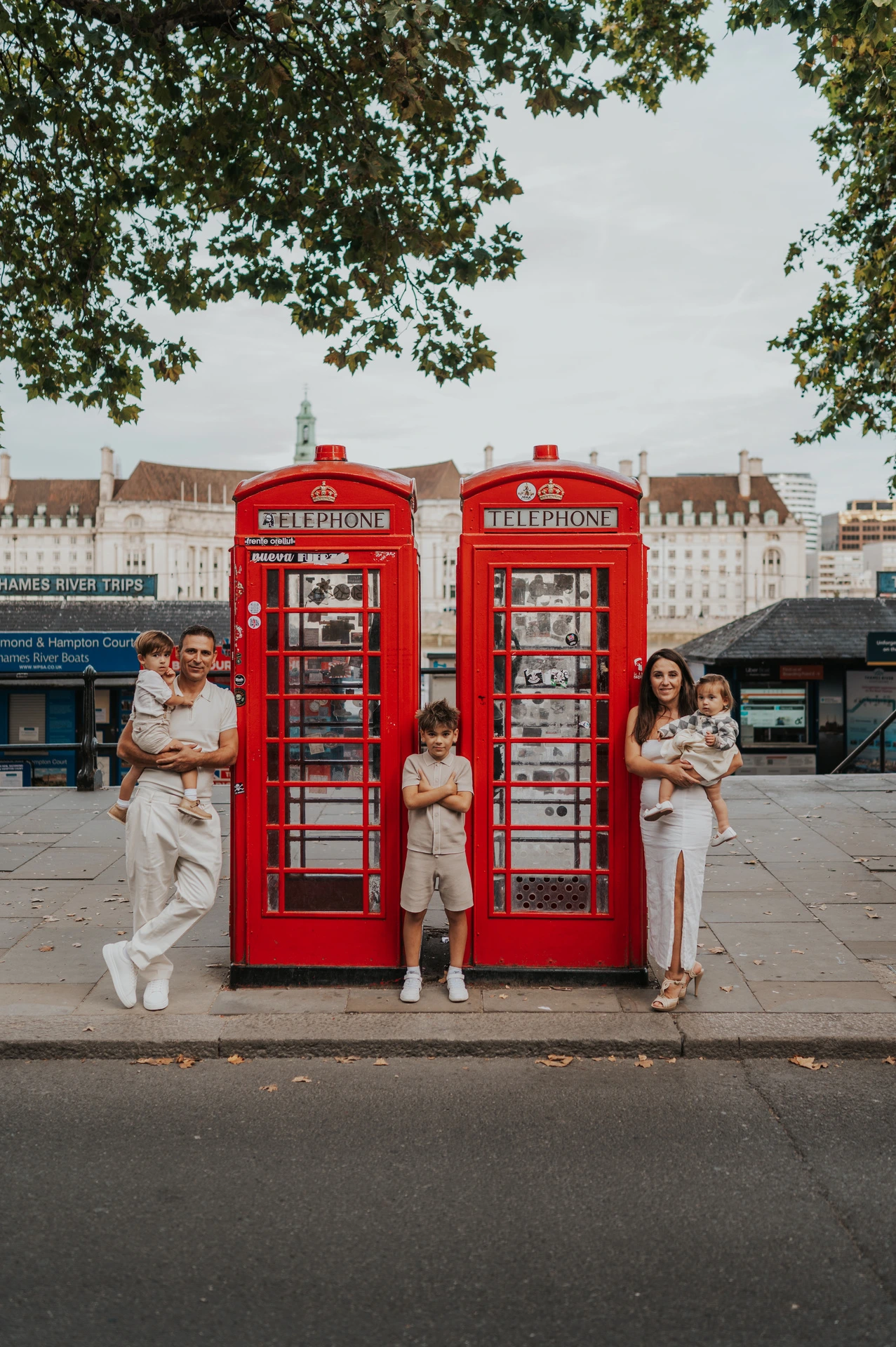 Wide view of a family beside two red telephone boxes near Westminster Pier with trees overhead during their London family photoshoot.