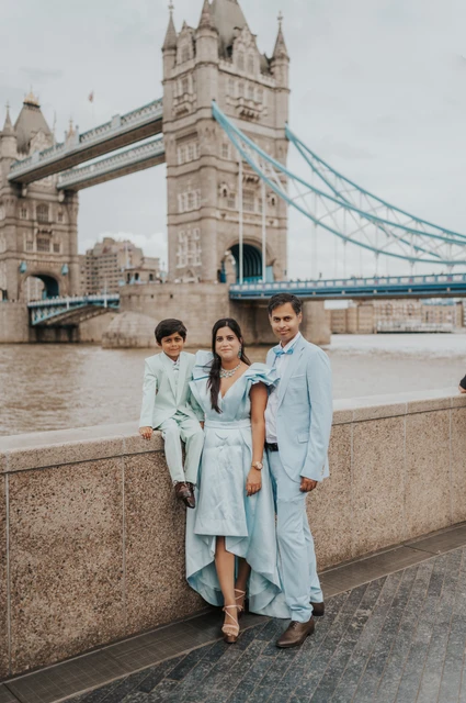 London family photoshoot: soft colours and the Tower Bridge view A family of three in soft blue and mint outfits beside the stone riverside wall with Tower Bridge behind during their London family photoshoot.