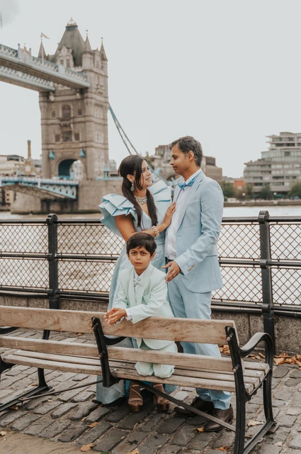 London family photoshoot: the family beside a wooden riverside bench A man and woman standing behind a child seated on a wooden bench beside the Thames and Tower Bridge during their London family photoshoot.