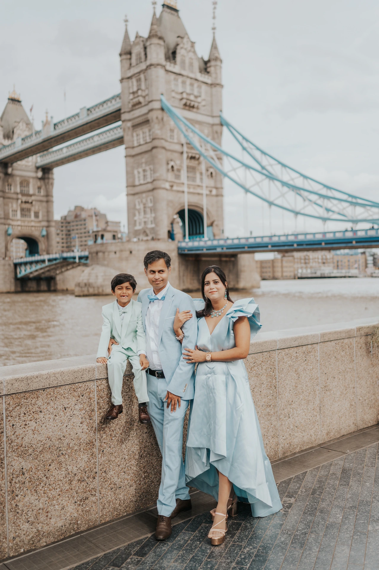 A family of three beside the Thames with a child sitting on the stone wall and Tower Bridge behind them during their London family photoshoot.