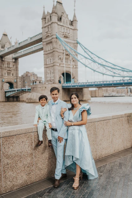 London family photoshoot: three together beside the Thames A family of three beside the Thames with a child sitting on the stone wall and Tower Bridge behind them during their London family photoshoot.