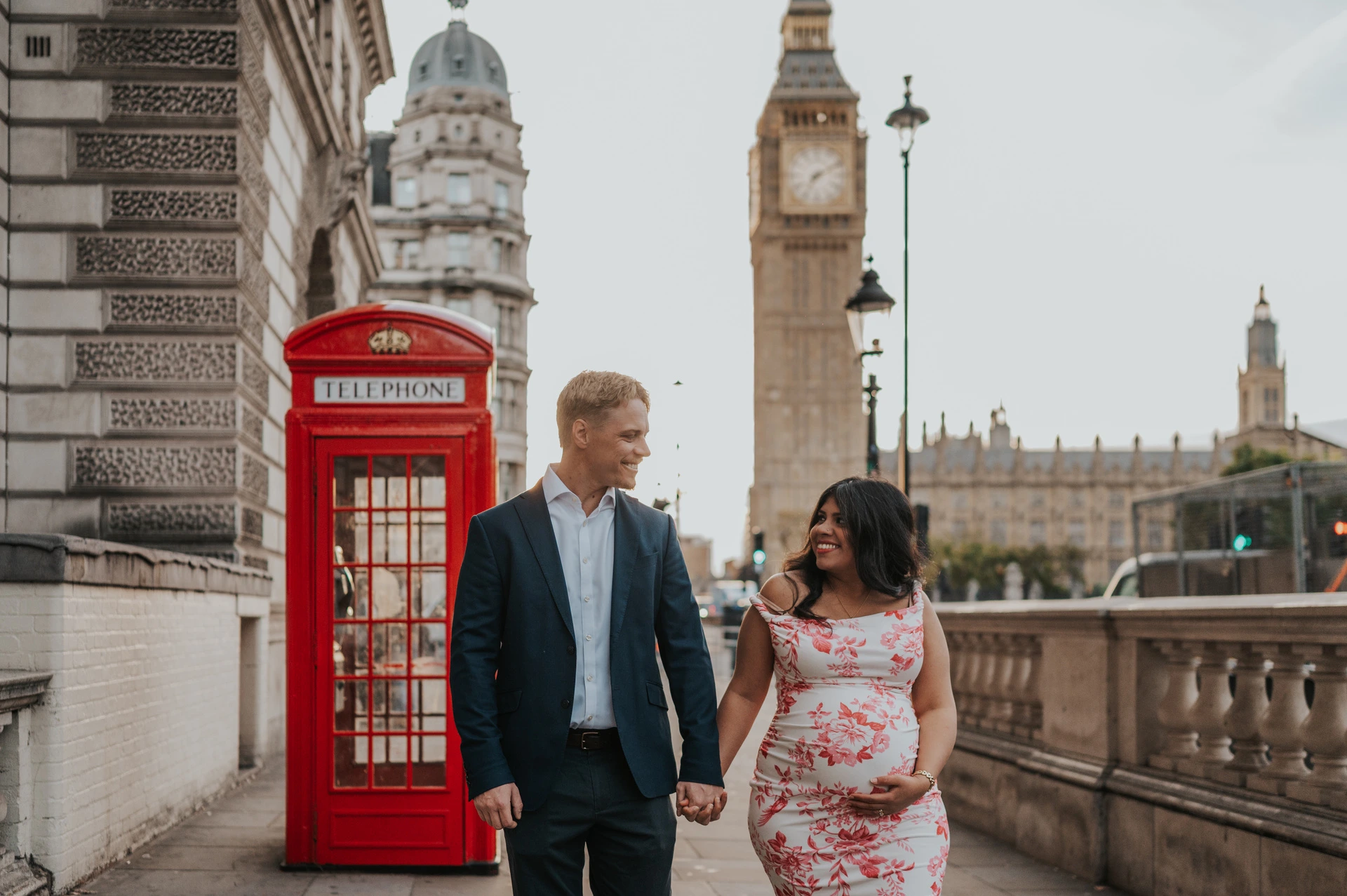 Landscape view of the couple smiling at one another beside a red telephone box, with Big Ben above them, during their London maternity photoshoot.