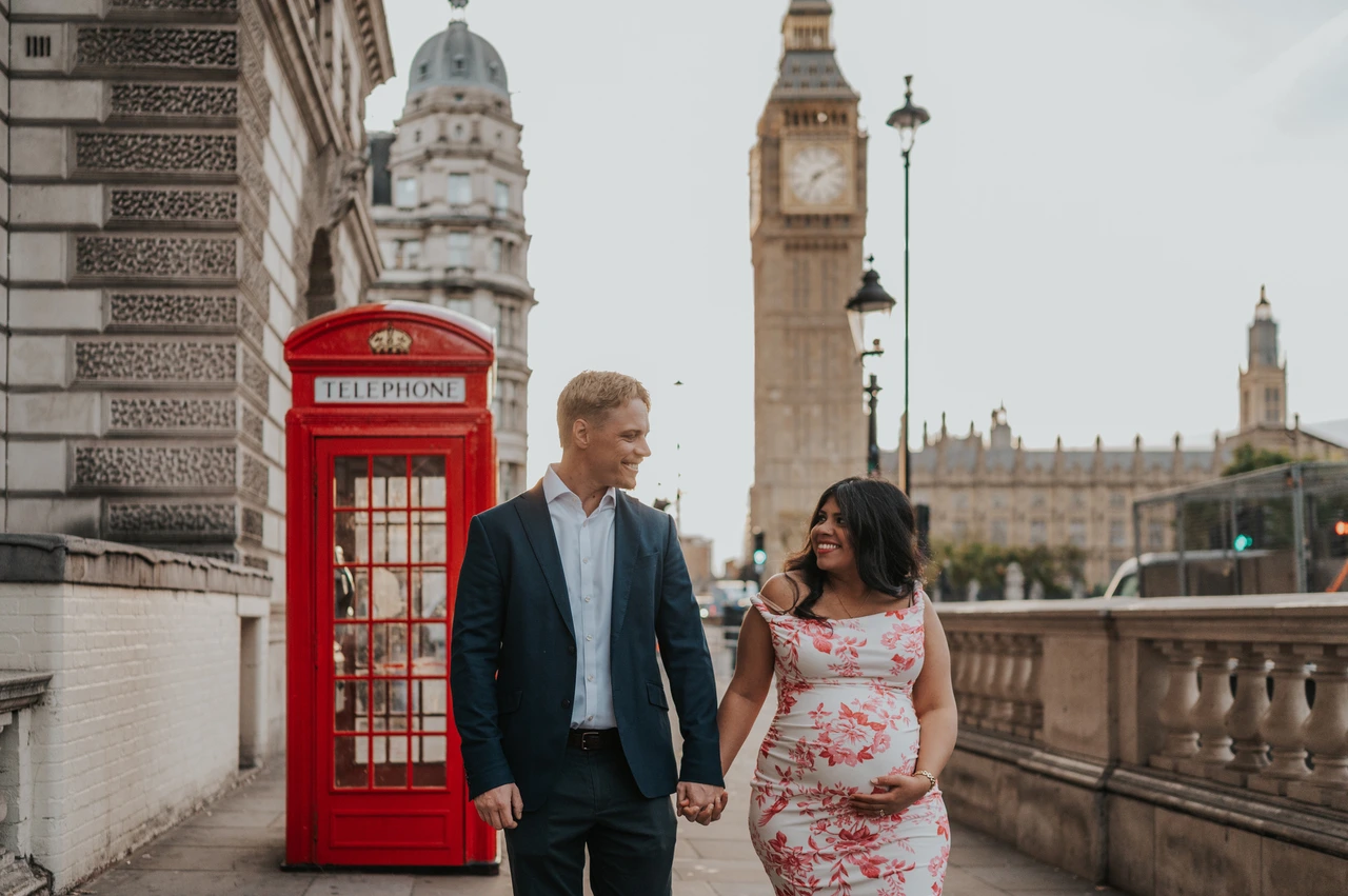 A wide Big Ben view for a London maternity photoshoot Landscape view of the couple smiling at one another beside a red telephone box, with Big Ben above them, during their London maternity photoshoot.