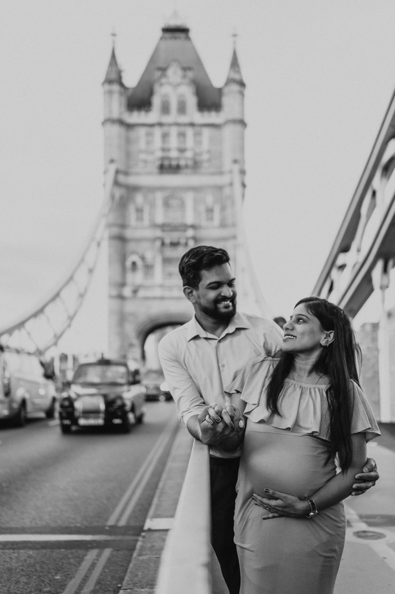 In black and white, a couple look towards each other beside Tower Bridge’s blue railing, with her hand resting on her bump and a bridge tower behind during their London maternity photoshoot.