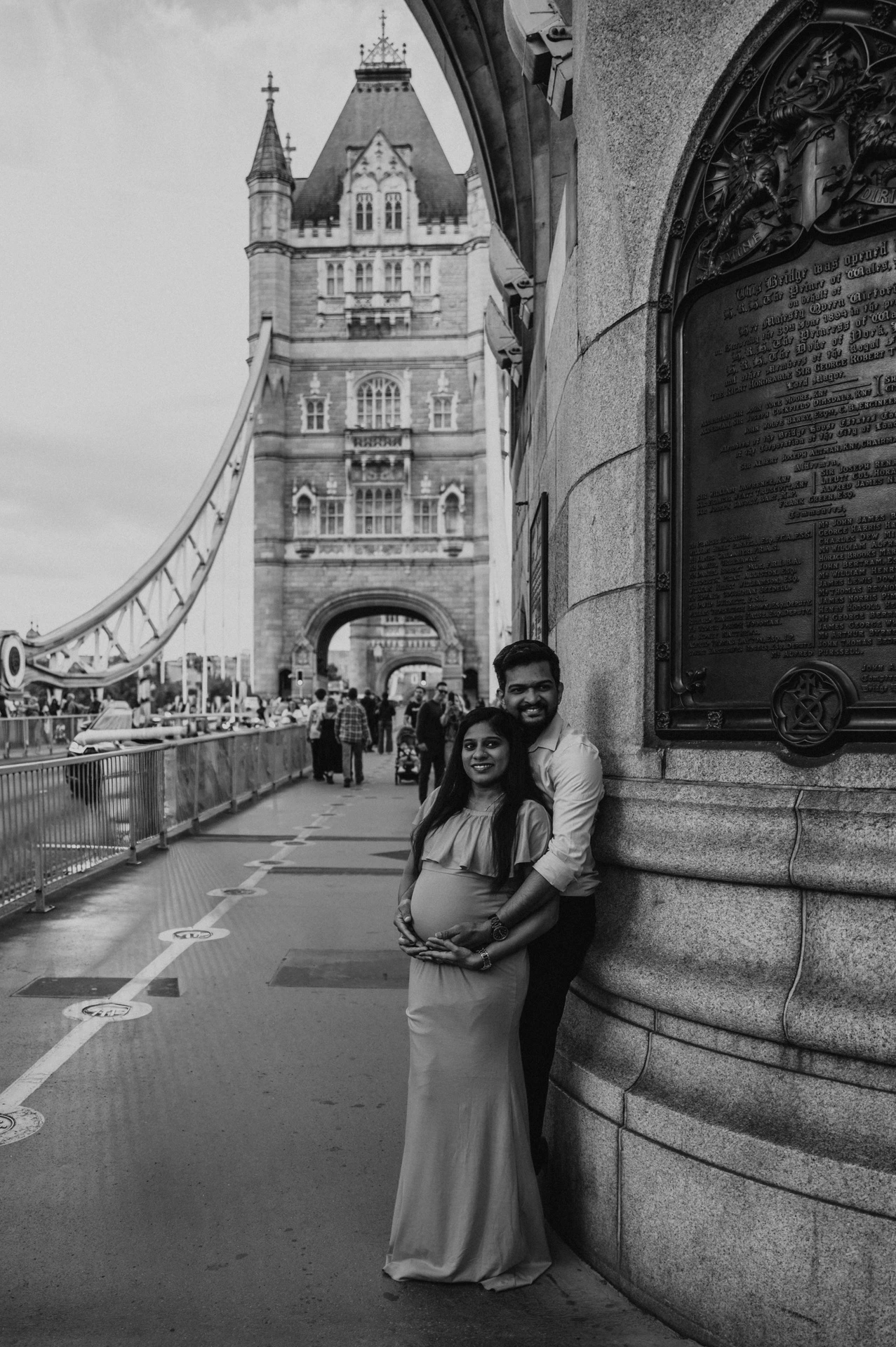 A black-and-white portrait shows a couple together beside Tower Bridge’s stone wall and plaque, with a bridge tower behind during their London maternity photoshoot.