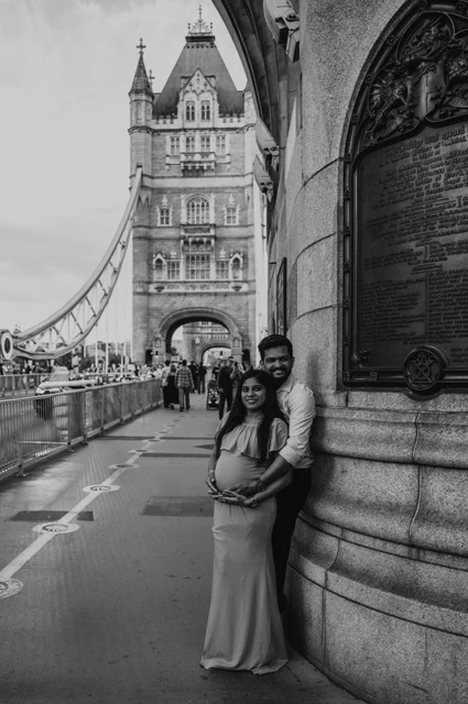 London maternity photoshoot: black-and-white portrait beside the bridge stonework. A black-and-white portrait shows a couple together beside Tower Bridge’s stone wall and plaque, with a bridge tower behind during their London maternity photoshoot.