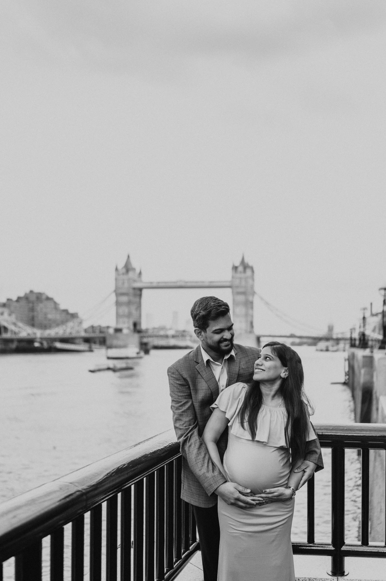 A black-and-white portrait shows a couple together at a dark riverside railing, with Tower Bridge across the Thames behind them during their London maternity photoshoot.