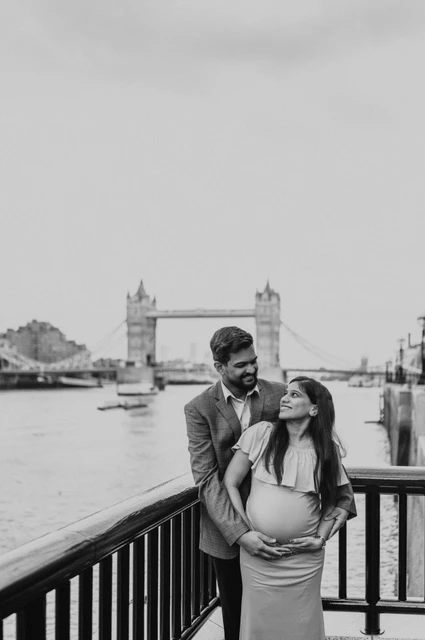 London maternity photoshoot: black-and-white couple portrait by the Thames. A black-and-white portrait shows a couple together at a dark riverside railing, with Tower Bridge across the Thames behind them during their London maternity photoshoot.