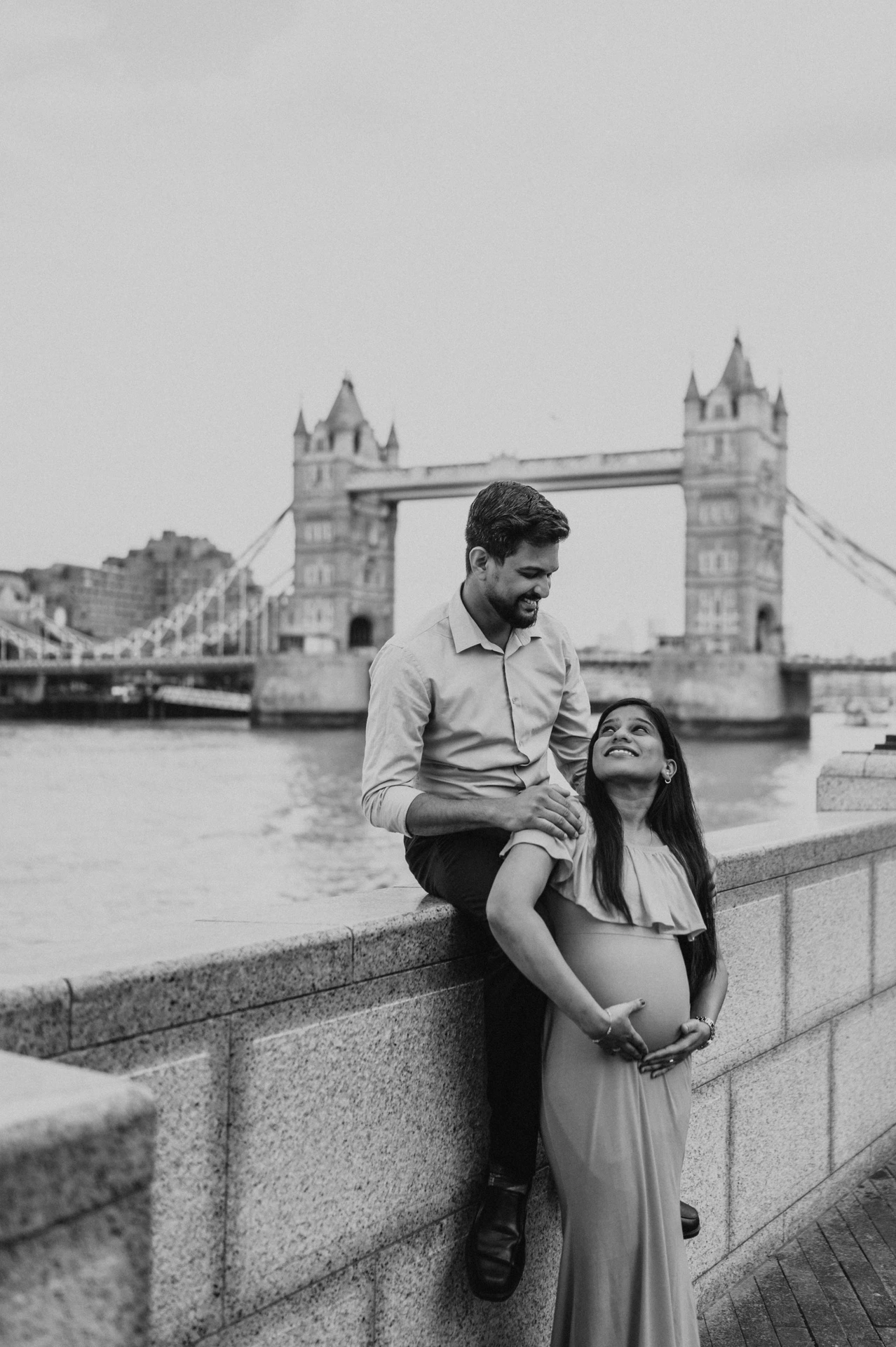 In black and white, a man sits on the stone river wall behind his pregnant partner, with Tower Bridge in the background during their London maternity photoshoot.