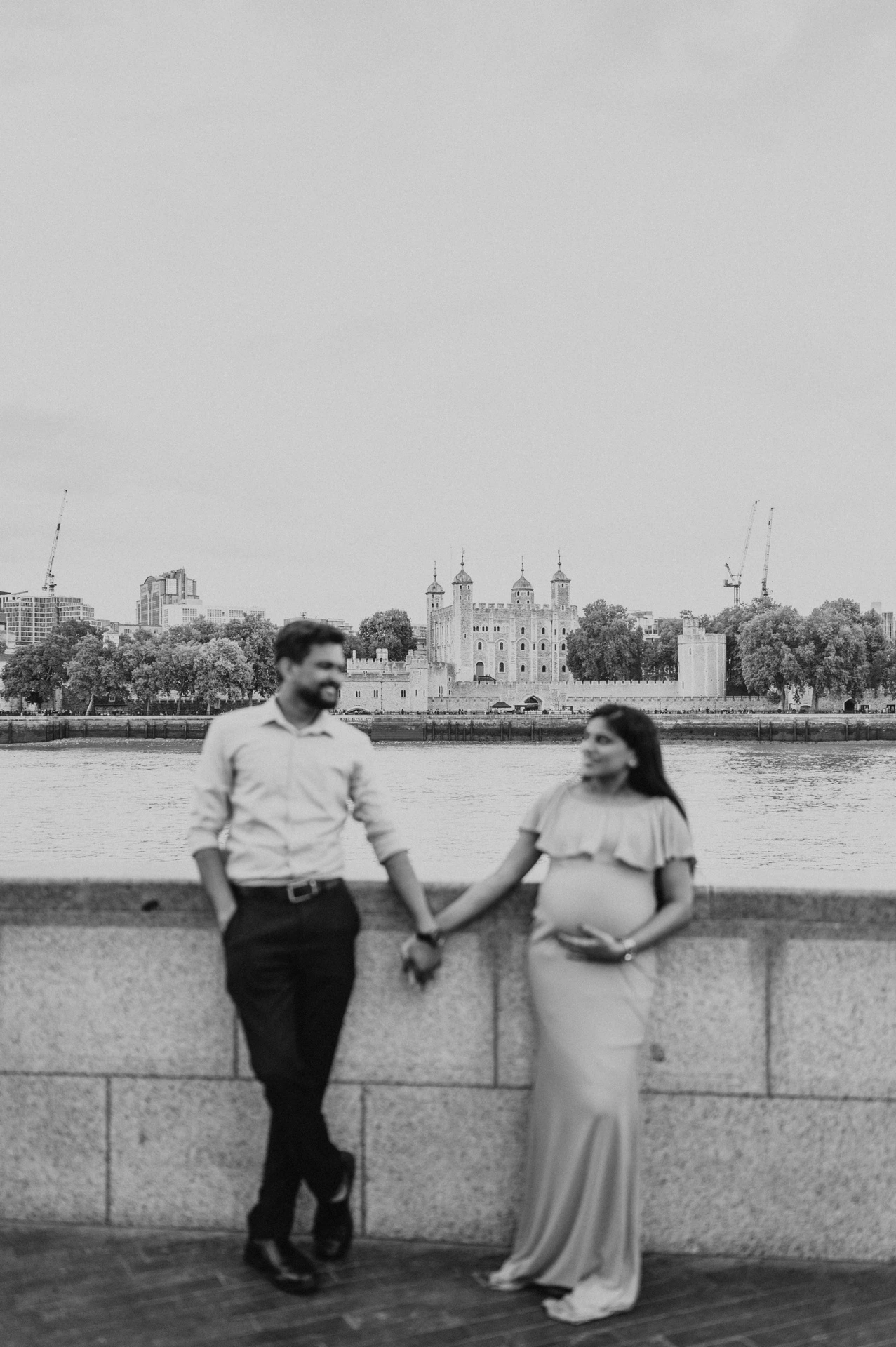 In black and white, a couple hold hands beside the river wall, with the Tower of London visible across the Thames during their London maternity photoshoot.