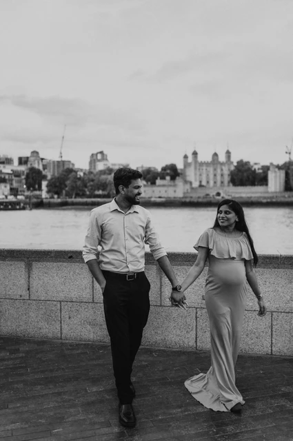 London maternity photoshoot: black-and-white walk along the river wall. In black and white, a couple walk hand in hand beside the Queen’s Walk river wall, with the woman’s long dress moving around her feet during their London maternity photoshoot.
