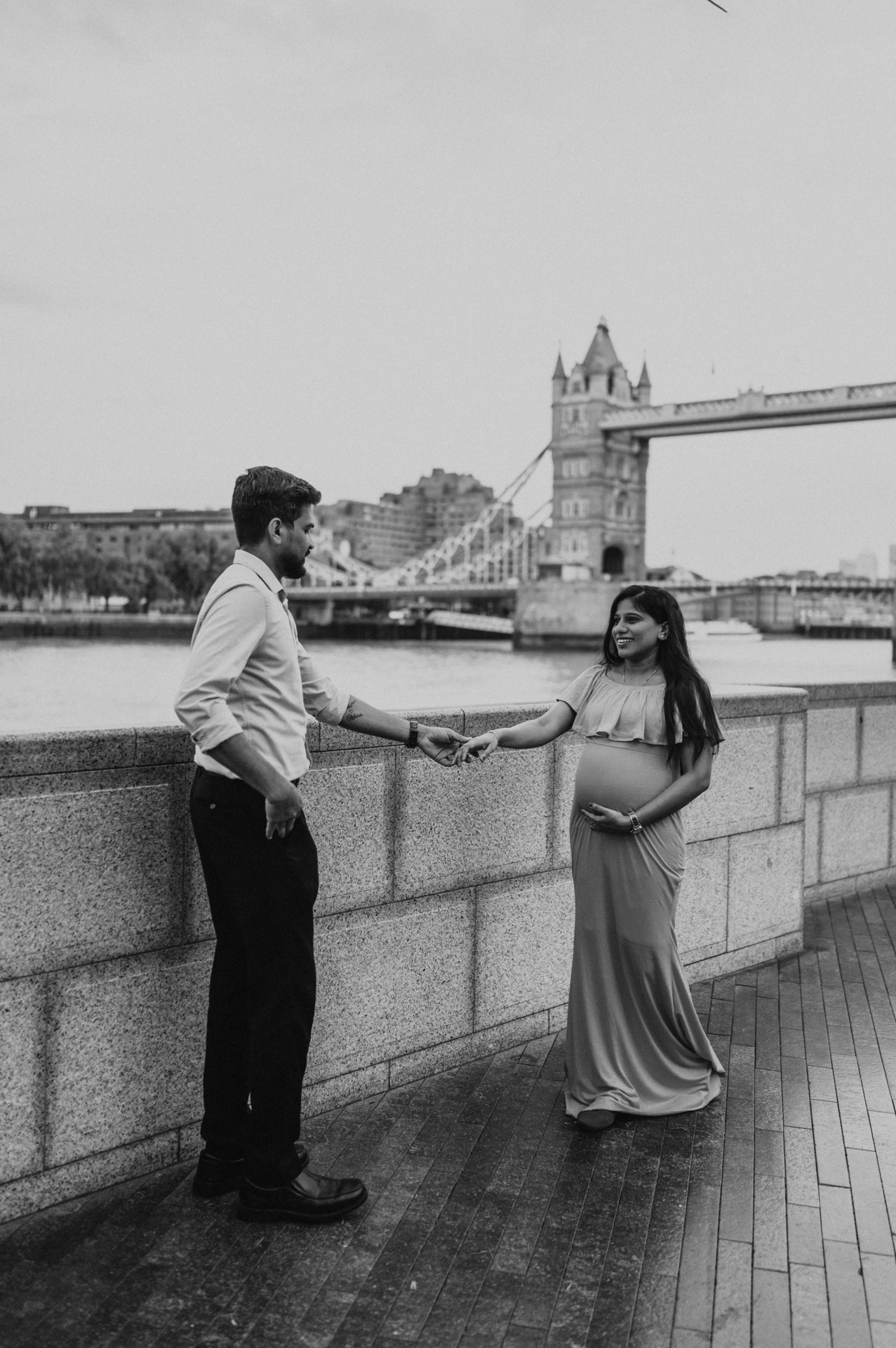 In black and white, a couple face each other and hold hands beside the stone river wall, with Tower Bridge behind during their London maternity photoshoot.