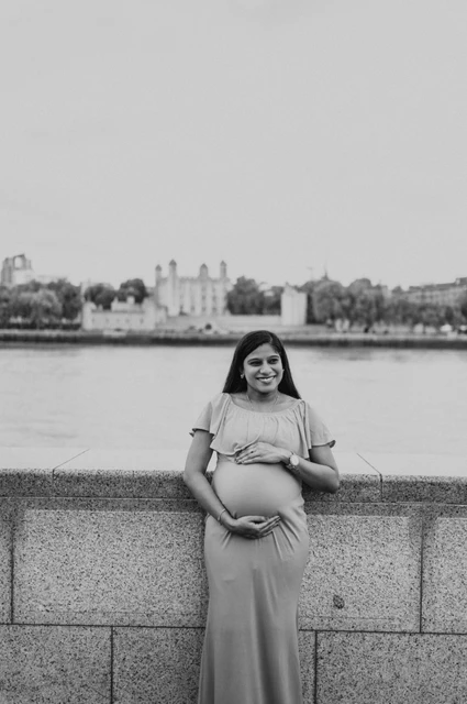 London maternity photoshoot: black-and-white portrait with the Tower of London. A black-and-white portrait shows a pregnant woman holding her bump beside the river wall, with the Tower of London across the Thames during their London maternity photoshoot.