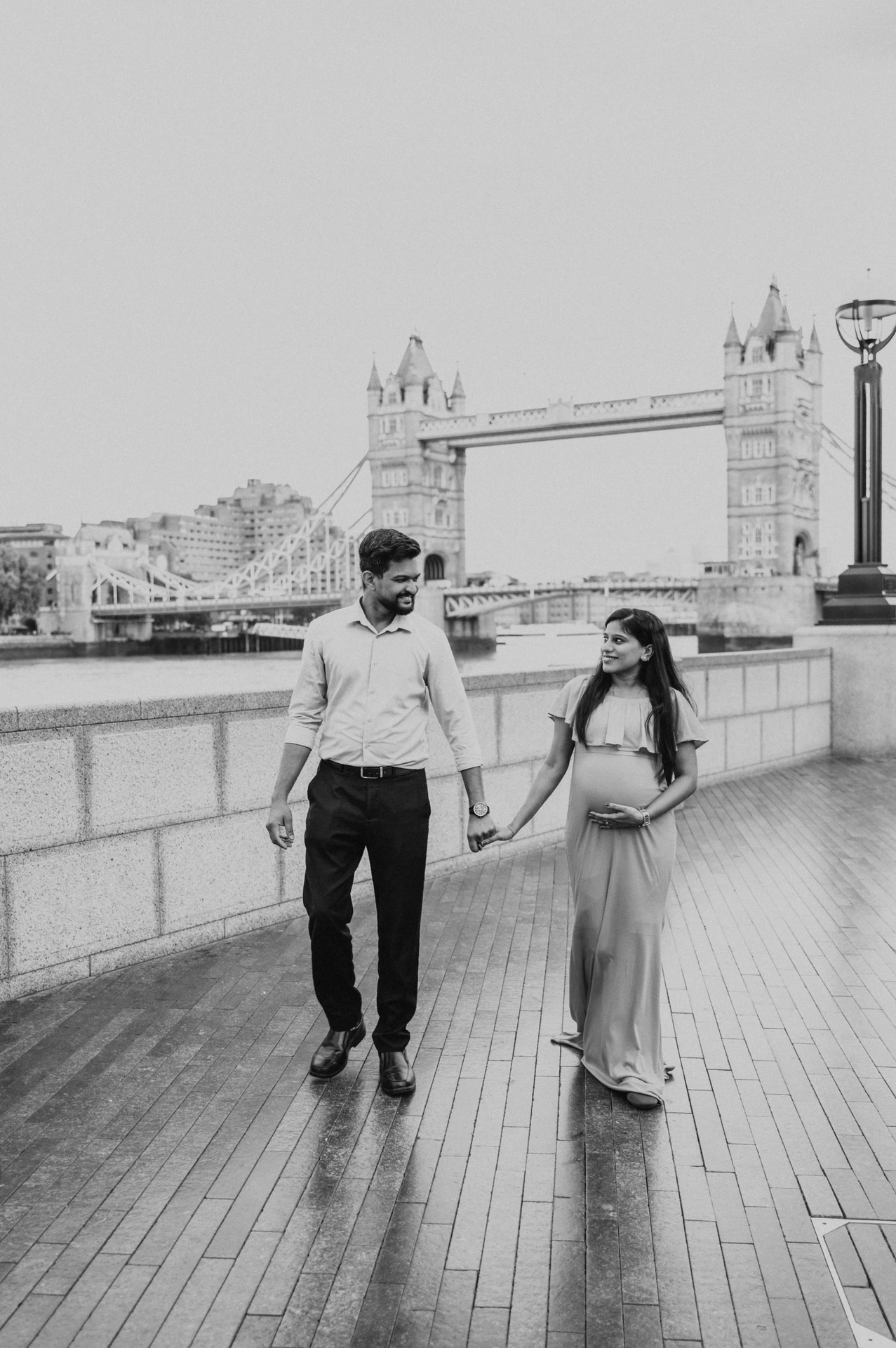 In black and white, a couple walk hand in hand beside the Queen’s Walk river wall, with Tower Bridge behind them during their London maternity photoshoot.