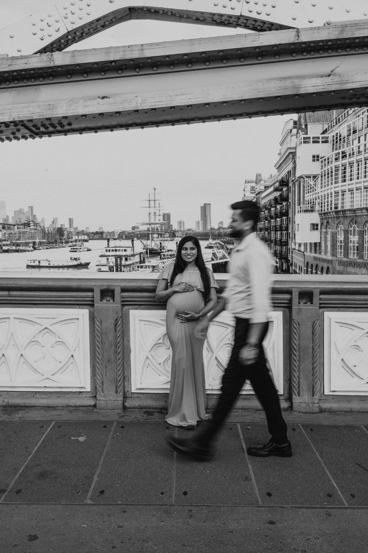 In black and white, a pregnant woman stands beside the Tower Bridge railing while her partner walks past, with the wharf buildings behind during their London maternity photoshoot.