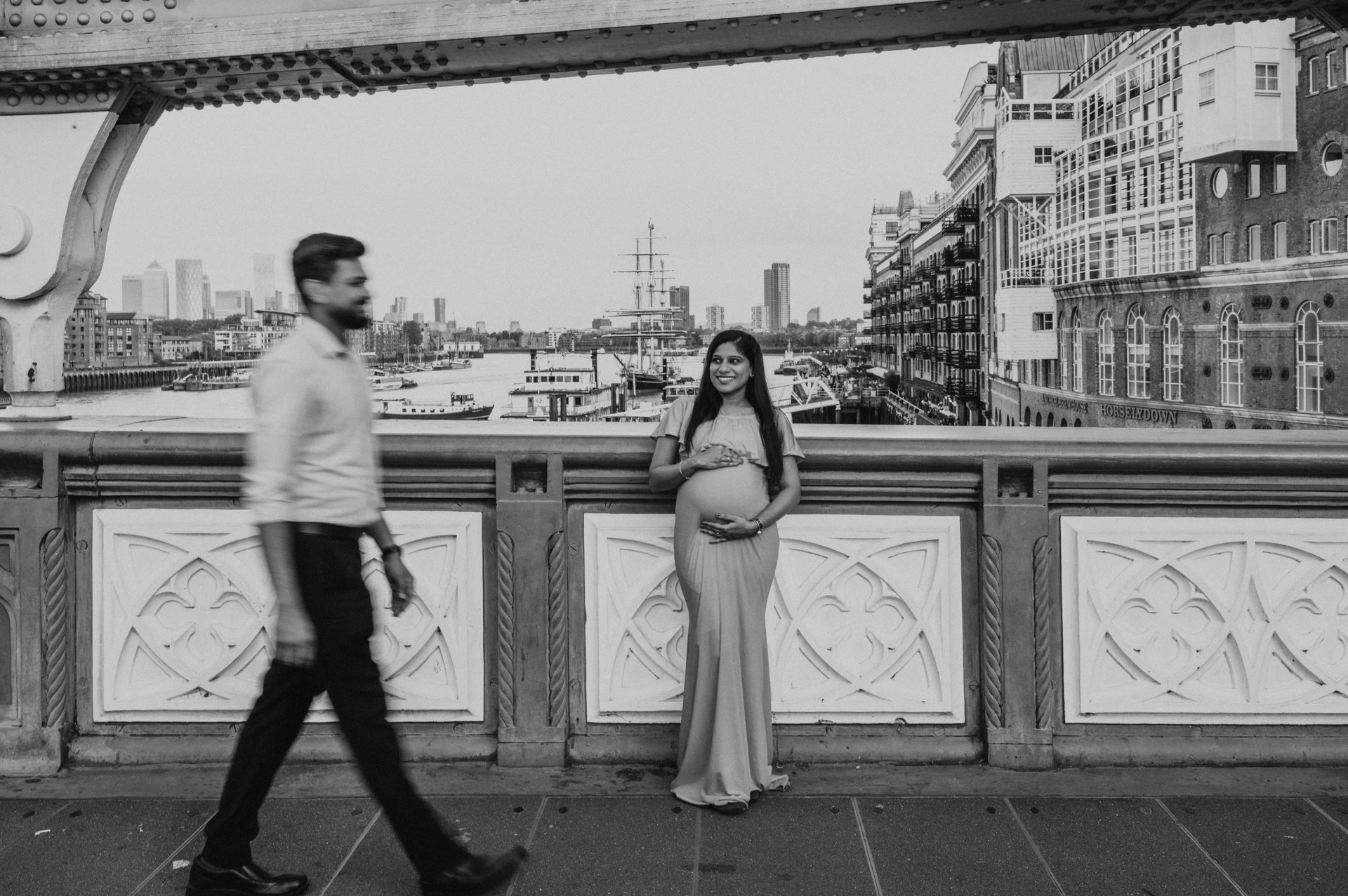 In a wide black-and-white photograph, a pregnant woman stands at the Tower Bridge railing while her partner walks in front of her, with riverside buildings behind during their London maternity photoshoot.