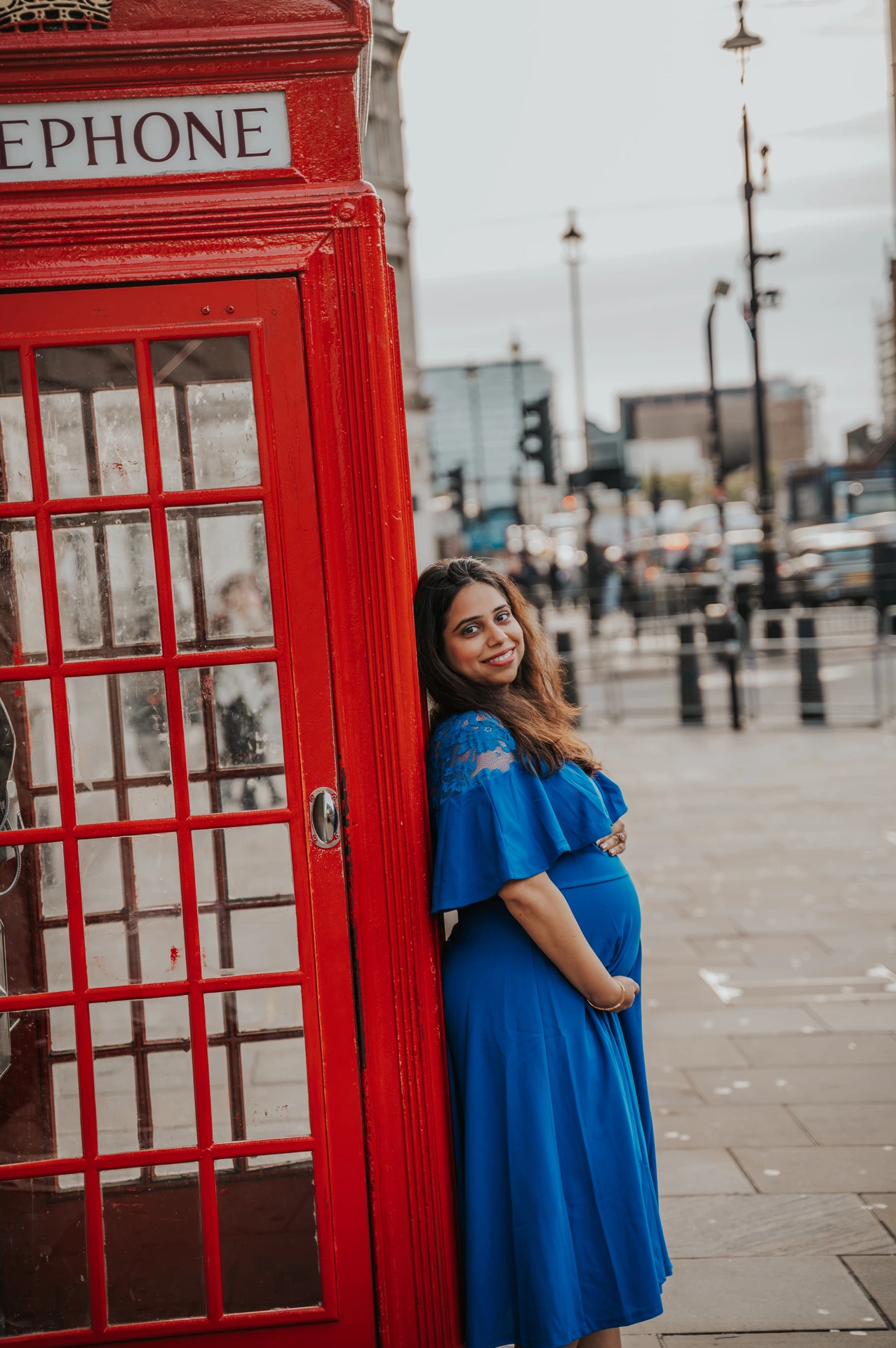 The expectant mother in a blue dress turns towards the camera beside a red telephone box during a London maternity photoshoot.