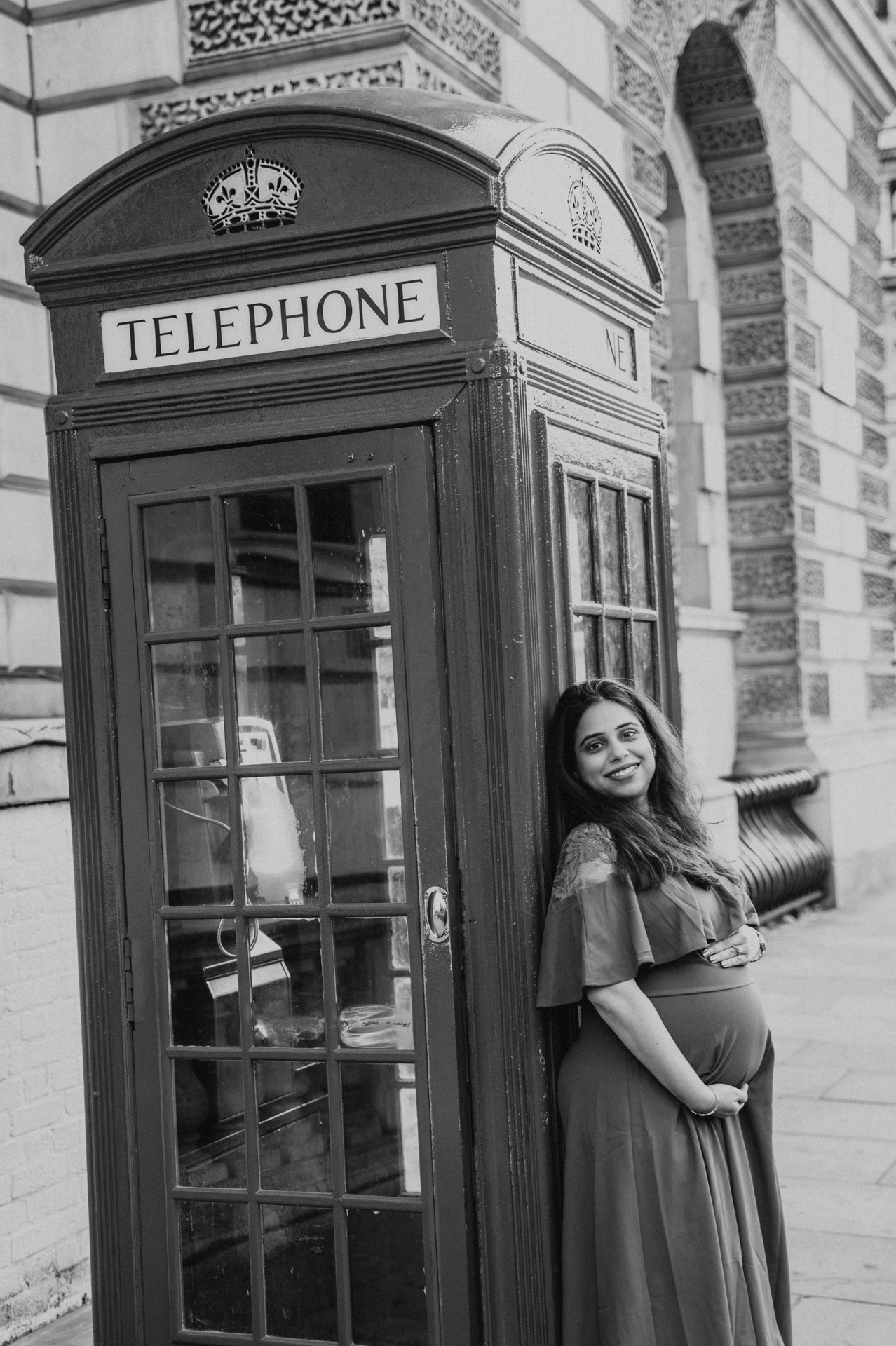 Black-and-white portrait of the expectant mother with her hands on her bump beside a telephone box during a London maternity photoshoot.