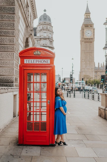London maternity photoshoot with a full-length blue dress portrait The expectant mother looks towards the camera beside a red telephone box with Big Ben in view during a London maternity photoshoot.