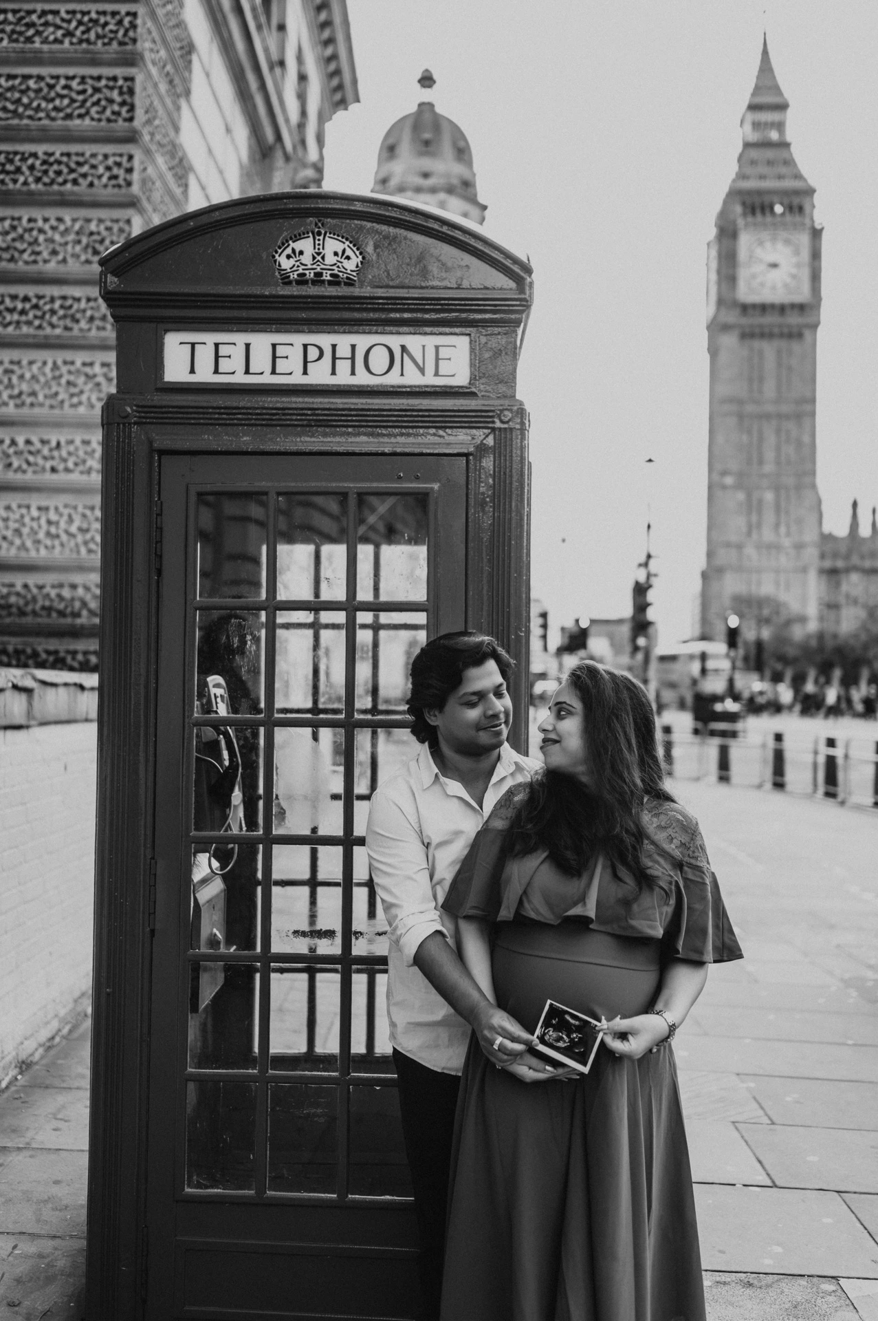 Black-and-white portrait of the couple holding an ultrasound scan beside a telephone box during a London maternity photoshoot.