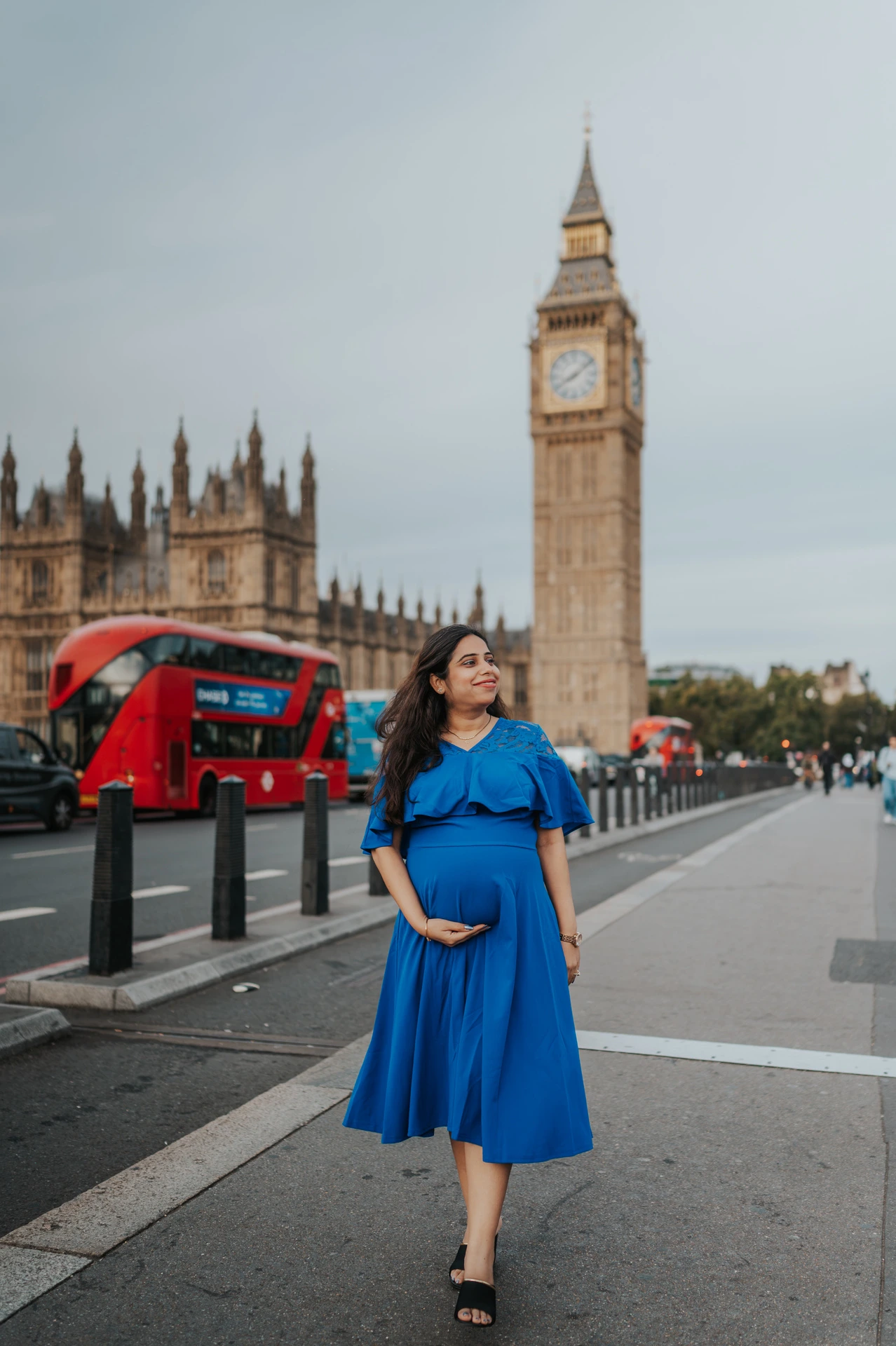 The expectant mother in a flowing blue dress looks to the side on Westminster Bridge during a London maternity photoshoot.