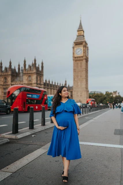 London maternity photoshoot with a full-length portrait on Westminster Bridge The expectant mother in a flowing blue dress looks to the side on Westminster Bridge during a London maternity photoshoot.