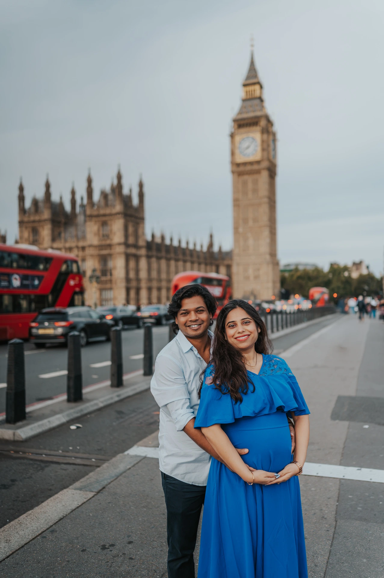 The couple smile towards the camera on Westminster Bridge with Big Ben and red buses behind them during a London maternity photoshoot.