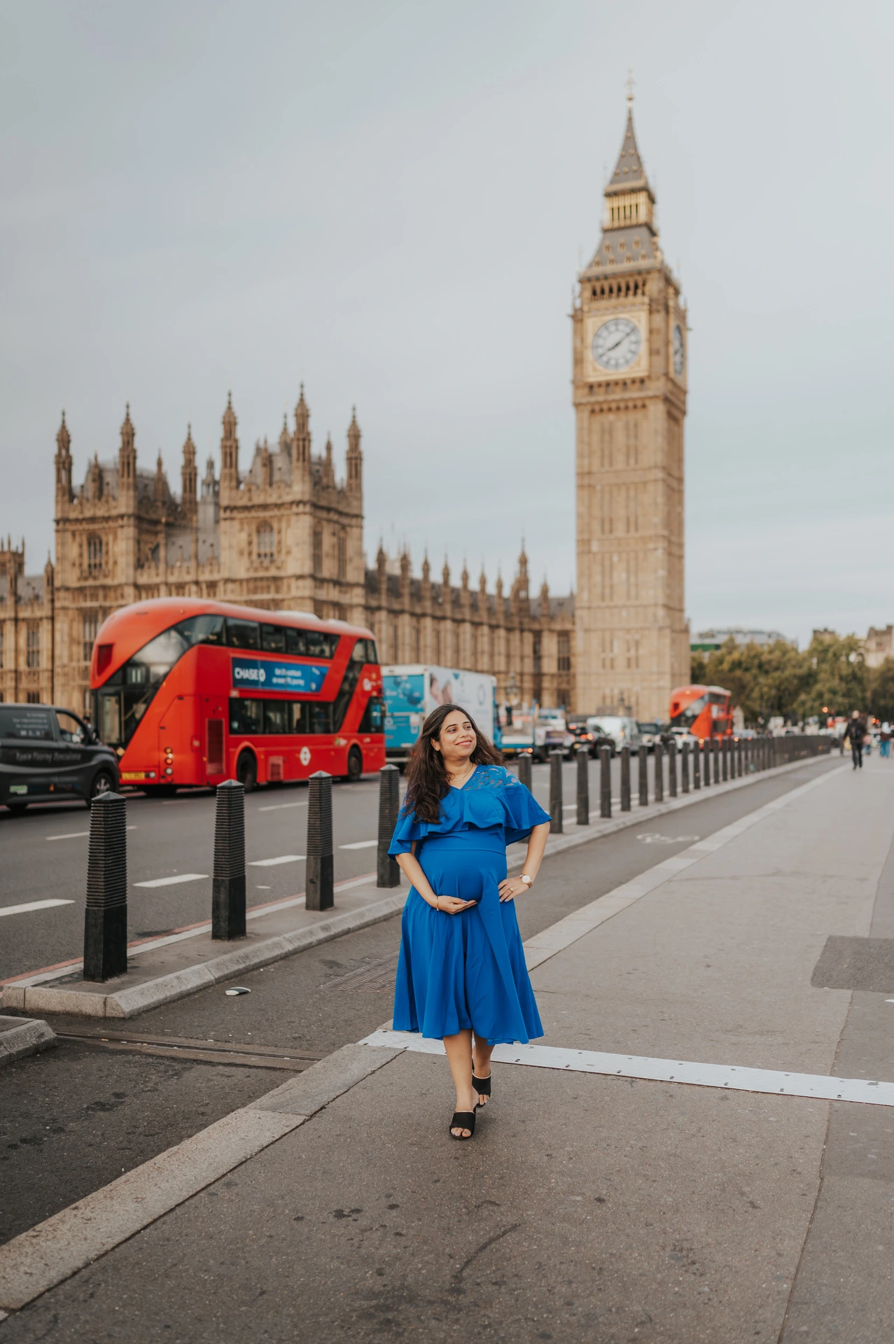 Wide full-length portrait of the expectant mother in a blue dress with Big Ben, Parliament and a red bus during a London maternity photoshoot.