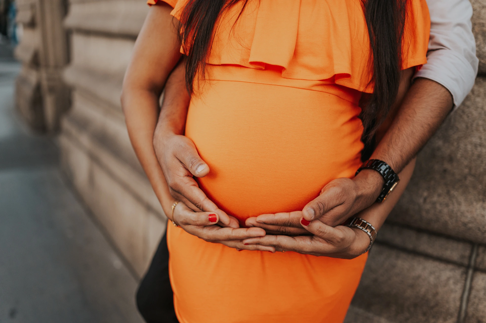 A close detail shows a couple’s hands together beneath her bump over an orange dress, beside the Tower Bridge stone wall during their London maternity photoshoot.