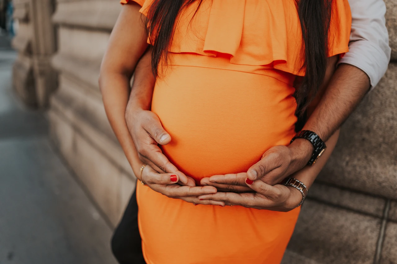 London maternity photoshoot: hands around her bump beside the bridge wall. A close detail shows a couple’s hands together beneath her bump over an orange dress, beside the Tower Bridge stone wall during their London maternity photoshoot.