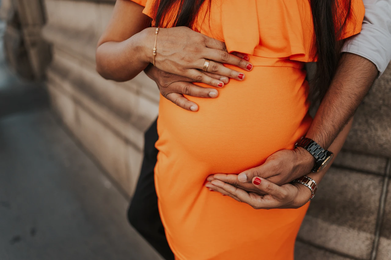 London maternity photoshoot: a close bump detail in the orange dress. A close detail shows one hand above her bump and the couple’s hands beneath it over an orange dress, beside the bridge stonework during their London maternity photoshoot.