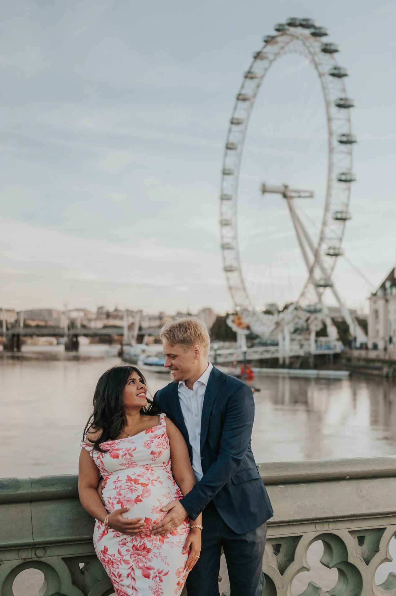 Close portrait of the couple smiling at each other above her bump, with the London Eye behind, during their London maternity photoshoot.