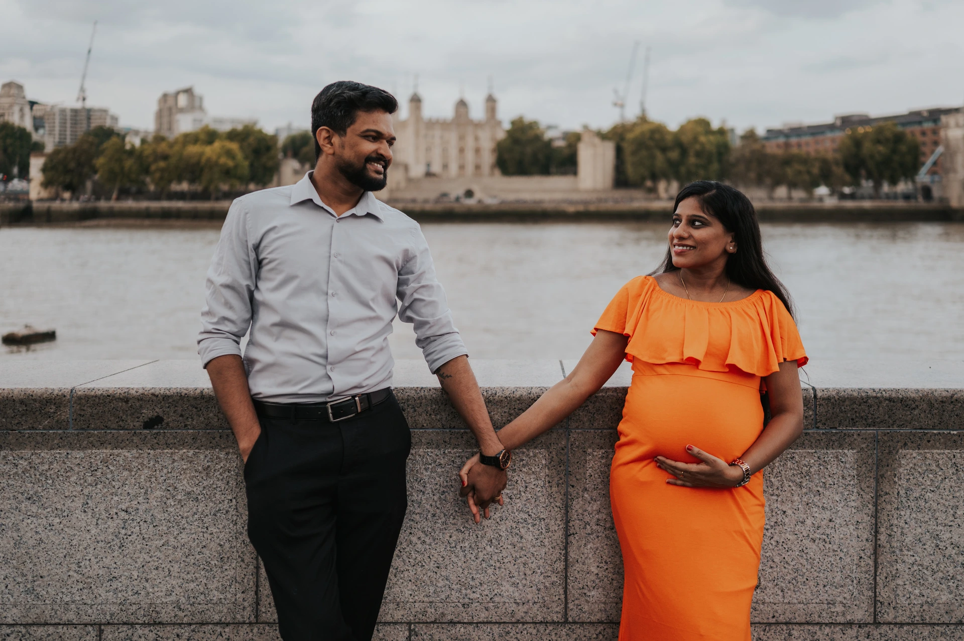 A close landscape portrait shows a couple holding hands beside the stone river wall, with her orange dress and the Tower of London beyond during their London maternity photoshoot.