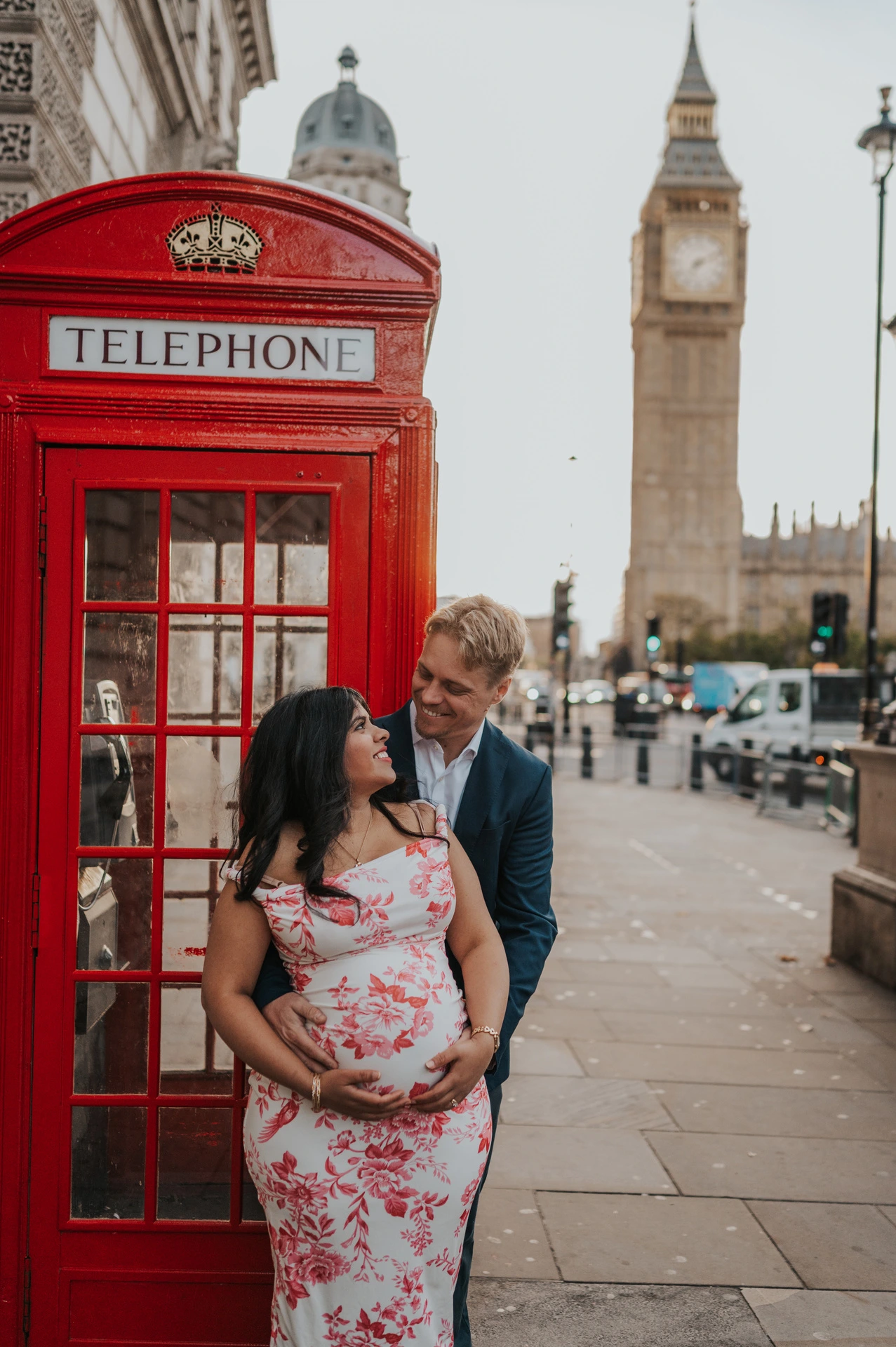 Close portrait of the couple looking at each other beside a red telephone box, with Big Ben behind, during their London maternity photoshoot.