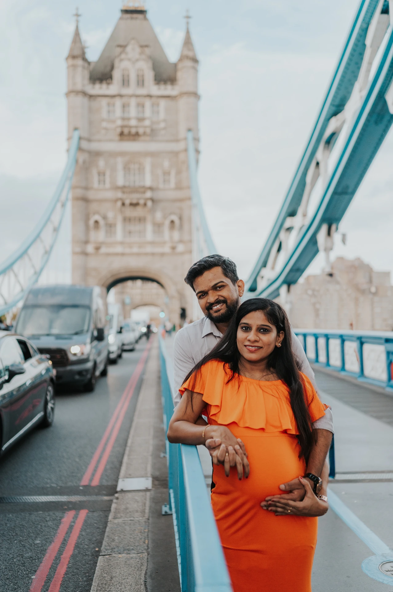 A couple stand together beside Tower Bridge’s blue railing, with the woman holding her bump in an orange dress and a bridge tower behind during their London maternity photoshoot.