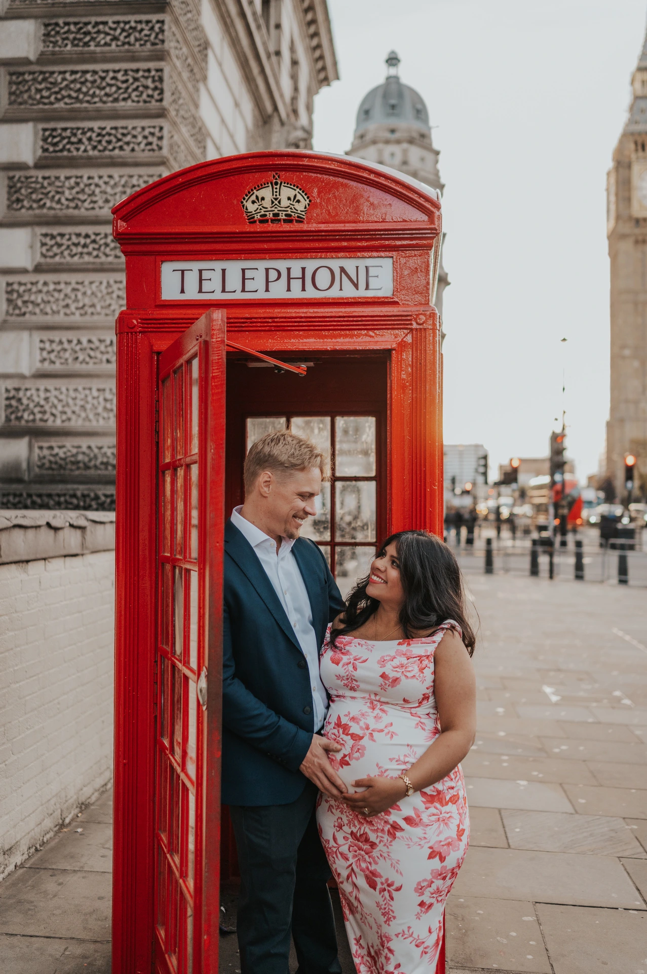 The couple looking at one another with their hands around her bump in a red telephone box doorway during their London maternity photoshoot.
