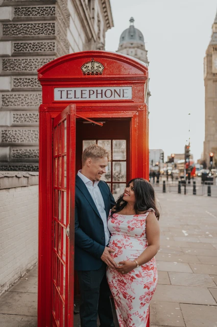 London maternity photoshoot with a couple in the telephone box doorway The couple looking at one another with their hands around her bump in a red telephone box doorway during their London maternity photoshoot.