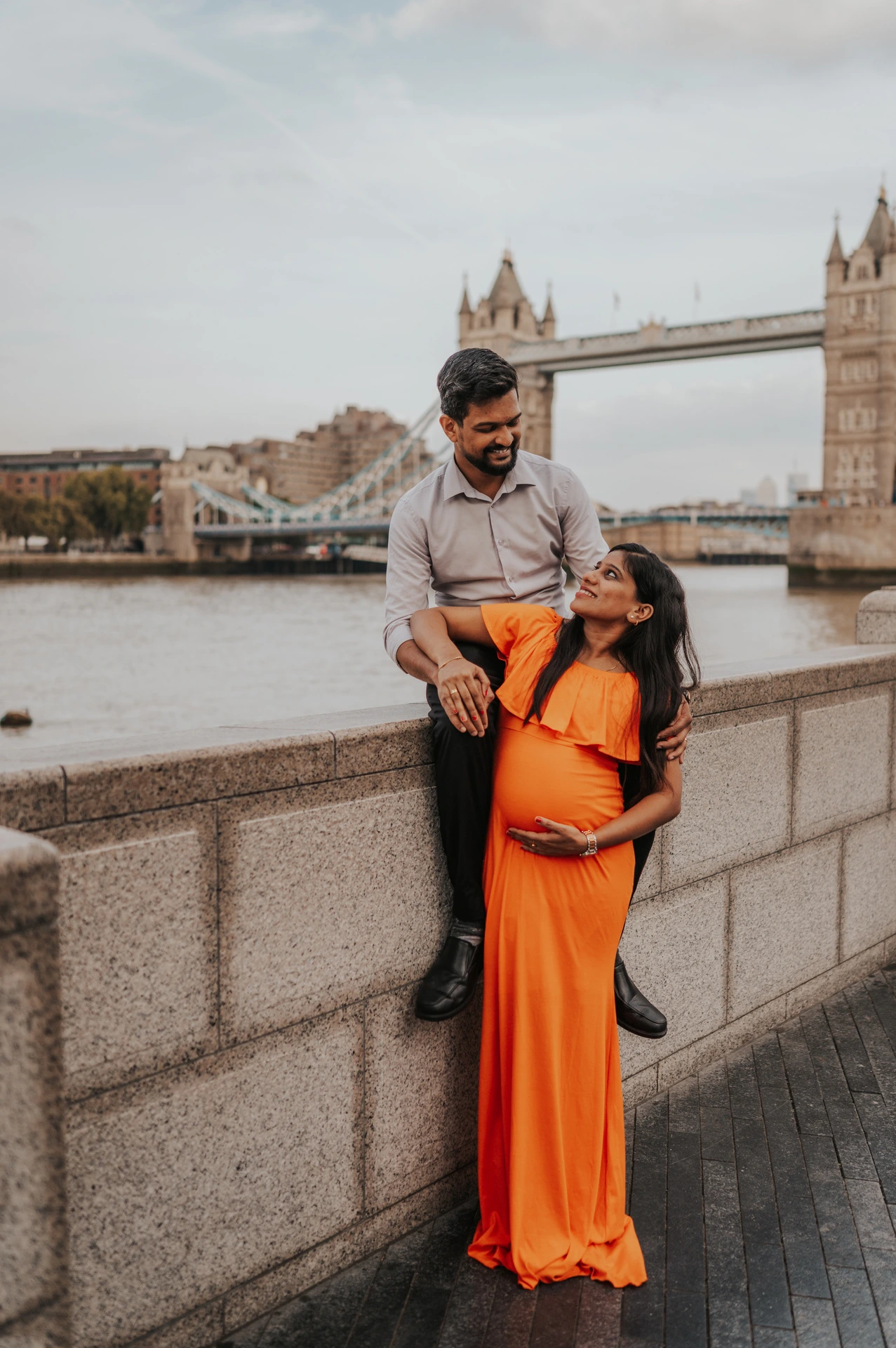 A pregnant woman in an orange dress reaches towards her partner seated on the river wall, with Tower Bridge across the Thames during their London maternity photoshoot.