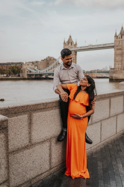 London maternity photoshoot: a close moment beside the Thames wall. A pregnant woman in an orange dress reaches towards her partner seated on the river wall, with Tower Bridge across the Thames during their London maternity photoshoot.