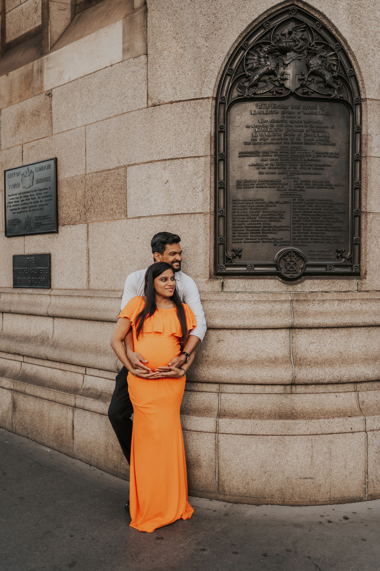 A couple look towards each other beside Tower Bridge’s curved stone wall and decorative plaque, with their hands around her bump during their London maternity photoshoot.