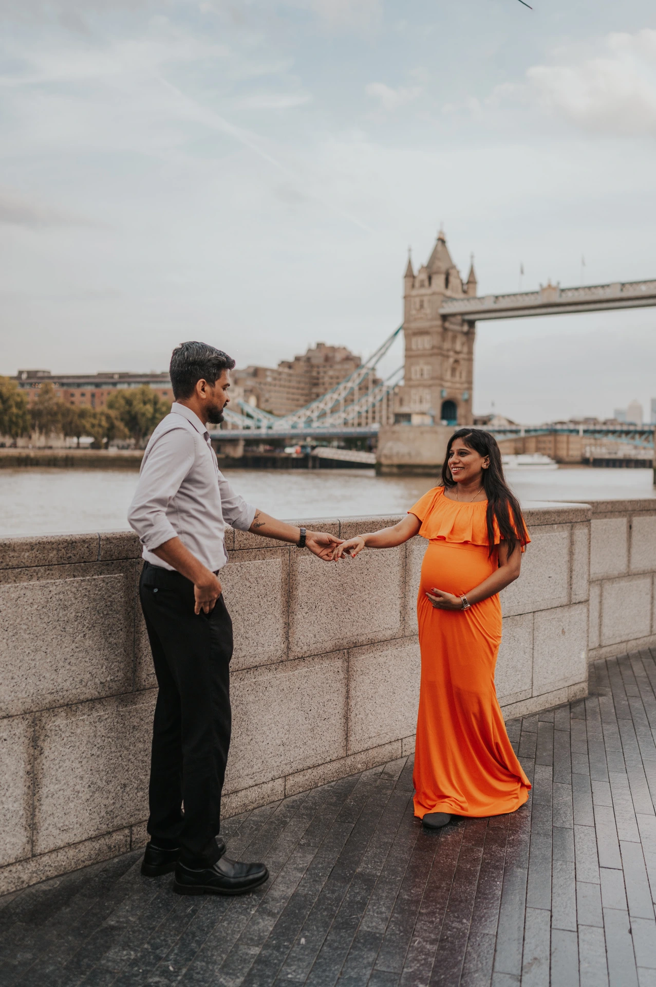 A couple hold hands while facing each other beside the Queen’s Walk river wall, with her orange dress and Tower Bridge in view during their London maternity photoshoot.
