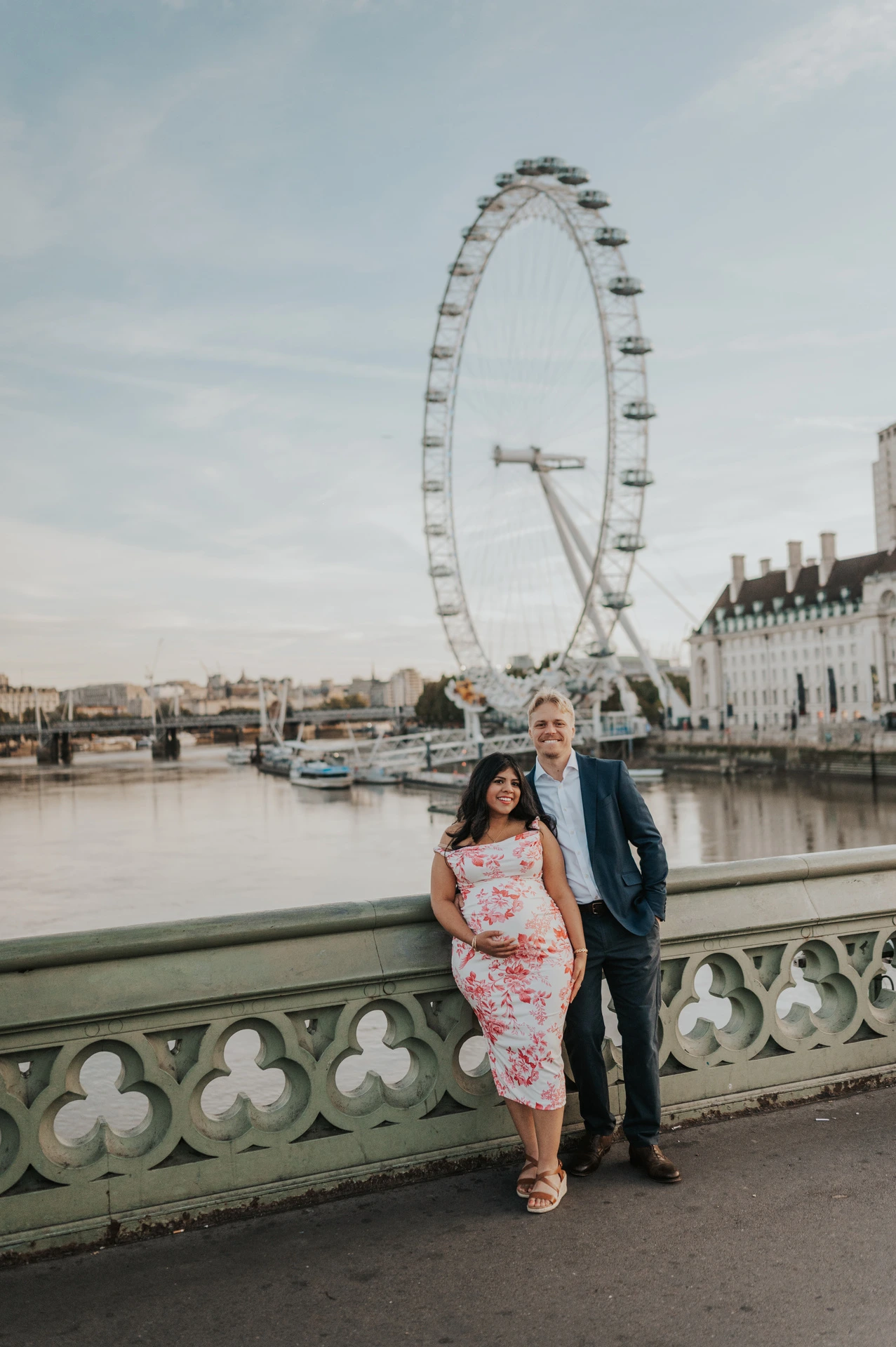 The couple standing beside the green Westminster Bridge railing with the London Eye across the Thames during their London maternity photoshoot.