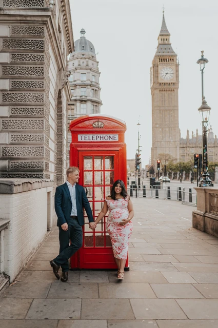 London maternity photoshoot with a classic red telephone box The couple standing hand in hand across the front of a red telephone box beneath Big Ben during their London maternity photoshoot.