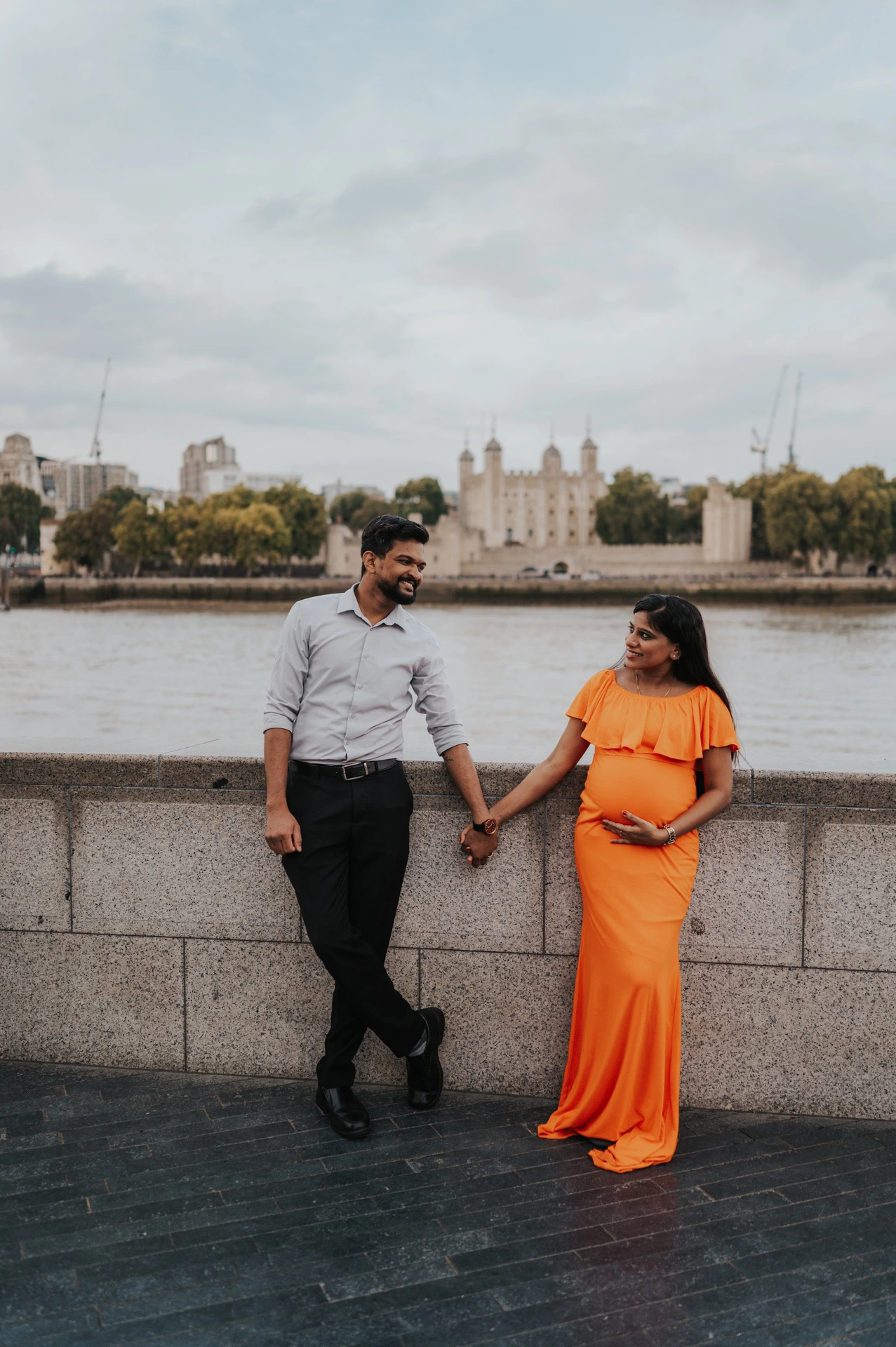 A couple hold hands beside the Queen’s Walk river wall, with the woman in an orange dress and the Tower of London across the Thames during their London maternity photoshoot.